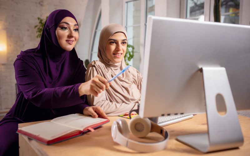 Pointing. Happy and young two muslim women at home during lesson, studying near computer, online education. Culture, traditions, modern people. Watching at PC monitor, shopping or talking.